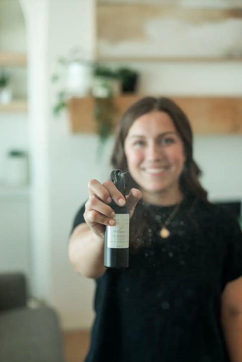 A smiling woman in a black shirt holds Shanti + Tula’s Natural Room & Linen Spray – Odor Neutralizer & Air Refresher, ready to demonstrate its use, with a blurred background of shelves and plants. Made in the USA.