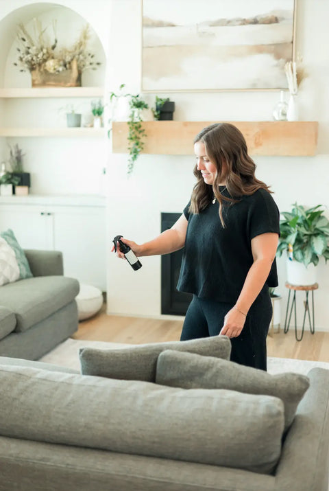 A woman in black clothes smiles as she uses Shanti + Tulas Natural Room & Linen Spray – Odor Neutralizer & Air Refresher on a gray couch in a bright, plant-filled living room with wooden shelves and light decor.