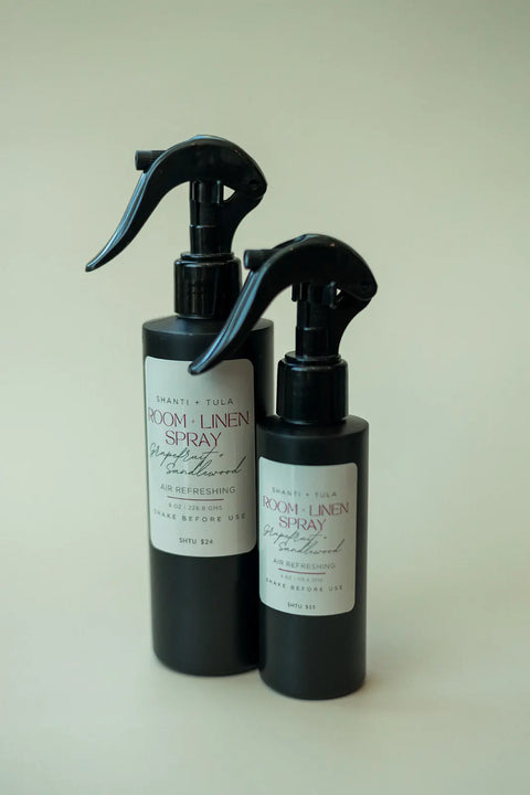 Two black Shanti + Tula Natural Room & Linen Spray bottles—odor neutralizers and air refreshers—stand side by side on a light surface. The white-labeled left bottle is taller, both made in the USA for freshening any space.