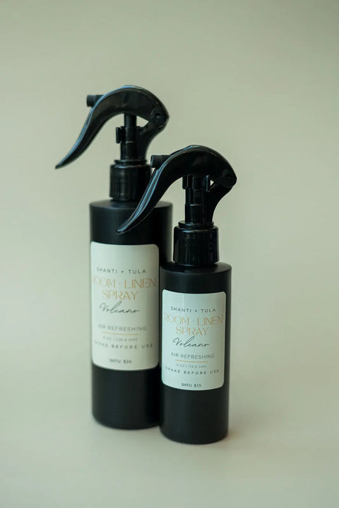 Two black spray bottles labeled “Natural Room & Linen Spray – Odor Neutralizer & Air Refresher” by Shanti + Tula stand on a beige surface, one tall and one short, both with black sprayers—perfect for freshening any room or linens.