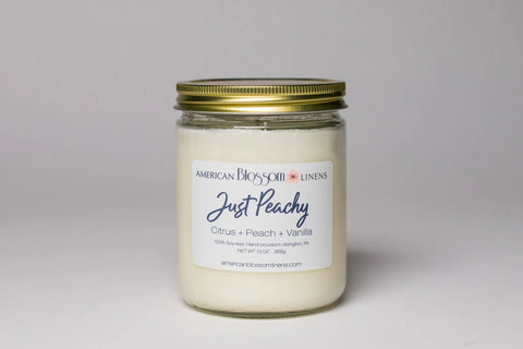The American Blossom Linens Hand Poured Soy Wax Scented Candle comes in a glass jar with a gold lid, labeled Just Peachy: Citrus + Peach + Vanilla. The plain white label lists the weight and maker’s website. Made by a local candle company.