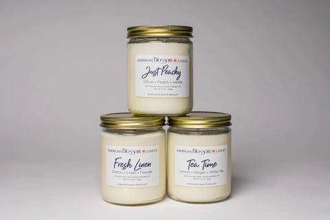 Three Hand Poured Soy Wax Scented Candles by American Blossom Linens with gold lids—“Just Peachy,” “Fresh Linen,” and “Tea Time”—are arranged in a pyramid against a white background.