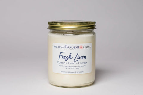 The Hand Poured Soy Wax Scented Candle by American Blossom Linens comes in a glass jar with a gold lid and Fresh Linen label, featuring a cotton, linen, and powder scent blend. The white candle sits against a simple light background.