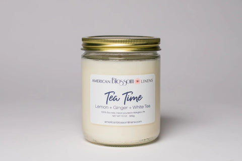 The Hand Poured Soy Wax Scented Candle by American Blossom Linens features a glass jar with gold lid and Tea Time: Lemon + Ginger + White Tea label, beautifully displayed on a plain white background.
