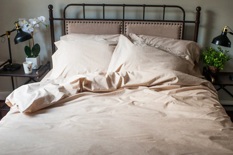 A neatly made bed with the American Blossom Linens Natural Colored Cotton Duvet Cover Set is set against a metal headboard. Nightstands hold black lamps, a potted orchid, a cup, and a small green plant on each side of the bed.