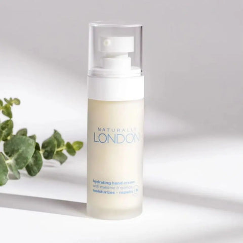 A bottle of Naturally London Hydrating Cream, 1.69oz, featuring a clear cap, white pump, and blue text stands on a white surface next to a small green sprig.
