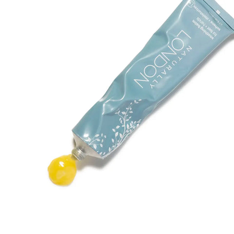 A blue tube labeled Naturally London Soothing Balm, 2.7oz, with a white leafy design dispenses thick yellow restorative moisturizer onto a white surface, inspired by Japanese skincare.