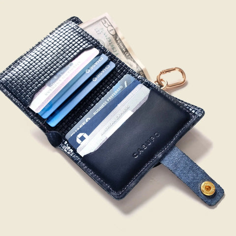 An open navy handcrafted wallet by CASUPO displays credit and ID cards, a folded U.S. dollar bill, and a gold keyring on a light beige background. It features a snap button closure and "CASUPO" embossed on the leather. Navy Limited Edition.