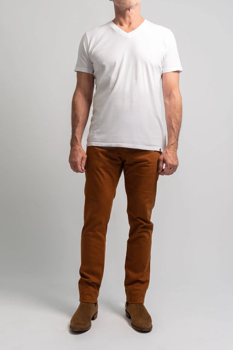 A man stands against a plain background wearing a white short-sleeve t-shirt, brown shoes, and Marc Nelson’s The Nelson Whiskey Stained Denim jeans. His face is not fully visible.
