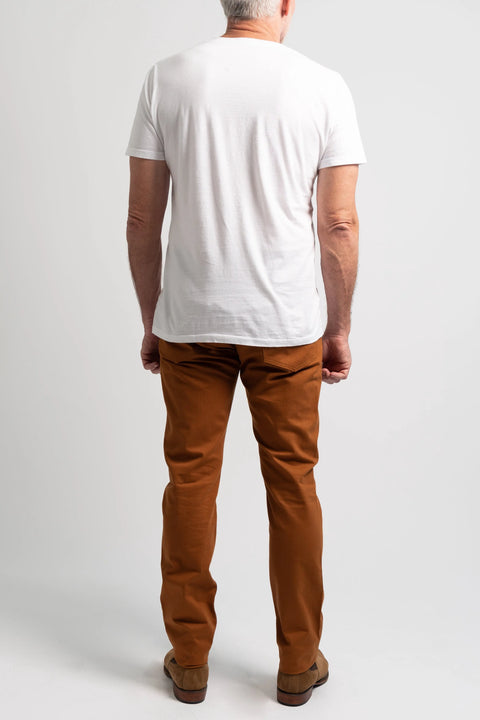 A man stands with his back to the camera, wearing a white T-shirt, brown shoes, and Marc Nelson’s The Nelson Whiskey Stained Denim jeans against a plain white background.
