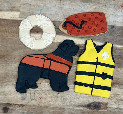 Celebrate Newfoundland Water Rescue heroes with Dogtown Bakerys Newfoundland Water Rescue Box—four artisan dog treats shaped like a life preserver, red rescue float, black dog in an orange vest, and yellow life jacket on a wooden surface.