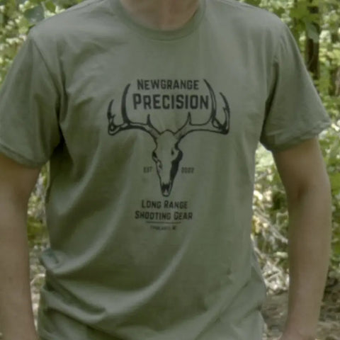 Someone wears an olive green American Lictor Collab Tee: Newgrange Logo, made from compacted ring-spun cotton with a deer skull graphic and “Est 2022, Long Range Shooting Gear, Tipalt, IL.” Leafy outdoors complete the scene.