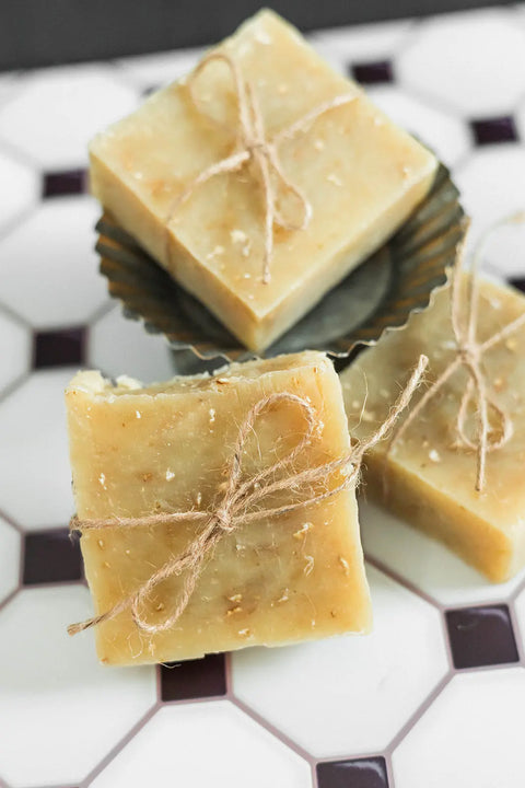 Three Shanti + Tula handmade soap bars, including the Oatmeal & Honey Body Bar—moisturizing, soothing, and USA-made—are tied with rustic twine and displayed on white tiles. One rests in a fluted metal dish, perfect for your skincare routine.