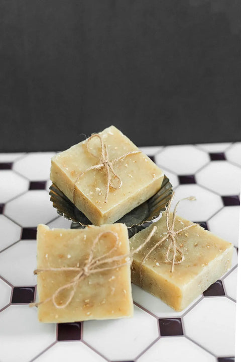 Three Shanti + Tula handmade soaps, including the Oatmeal & Honey Body Bar—moisturizing, soothing, and USA-made—feature a natural rustic look and are tied with twine on a white hexagonal tile, ideal for your skincare routine.