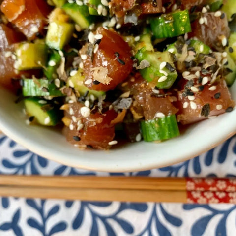 A close-up of a poke bowl topped with Oceans Balance Bonito Furikake, diced raw fish, cucumber, and sesame seeds in a white bowl, with wooden chopsticks on a blue and white patterned surface.