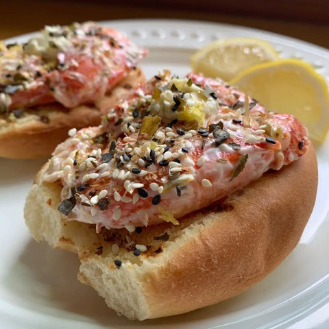 An open-faced sandwich on a bun with creamy seafood, sprinkled with Oceans Balance Bonito Furikake and regeneratively farmed seaweed, served with two lemon slices on a white plate.