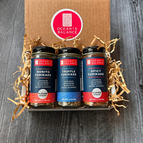 The Oceans Balance Furikake Gift Box, featuring Bonito, Truffle, and Spicy Japanese rice seasonings made with Maine seaweed, is displayed in a cardboard box with shredded paper on a dark wooden surface.
