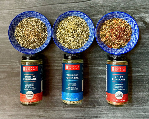Three bowls of Ocean’s Balance Furikake Assortment—Bonito, Truffle, and Spicy blends—are displayed above matching jars on a dark wooden surface, each featuring delicious seaweed in these unique Japanese rice seasonings.
