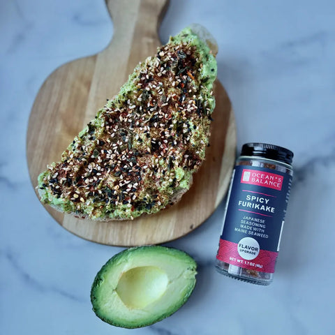 A slice of toast with avocado and Japanese rice seasoning sits on a wooden board beside an open jar of Ocean’s Balance Furikake Assortment and a halved avocado on a light surface.