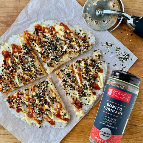 Four rice triangles topped with mayonnaise, sauce, and Japanese seasoning sit on parchment beside a pizza cutter. Nearby is a jar of Oceans Balance Furikake Assortment featuring its signature savory seaweed flavor.