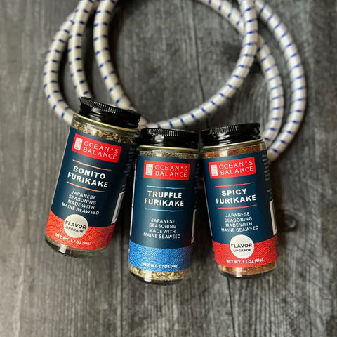 Oceans Balance Furikake Assortment—Bonito, Truffle, and Spicy—jars are displayed upright on dark wood with a coiled white and blue rope in the background, highlighting their flavorful seaweed and spice blends.