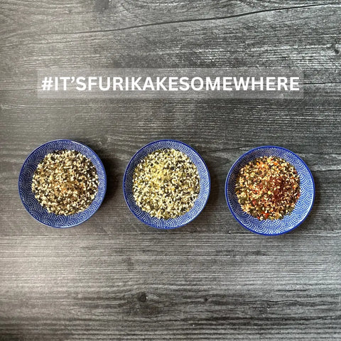 Three blue bowls with Oceans Balance Furikake Assortment, featuring Japanese rice seasonings and seaweed, are lined up on a dark wooden surface. The text above says #ITSFURIKAKESOMEWHERE.