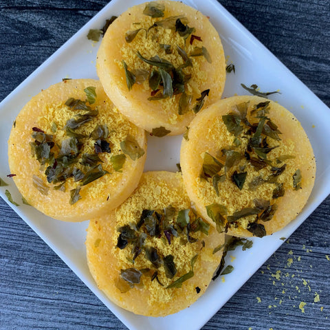 Four round pieces of yellow steamed cornbread topped with chopped greens and Oceans Balance Organic Sea Lettuce Flakes are arranged on a white square plate on a dark wooden surface.