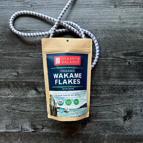 A brown resealable package of Oceans Balance Organic Wakame Flakes, certified USDA Organic, rests on a dark wooden surface with a blue and white rope behind it and coastal imagery on the package.