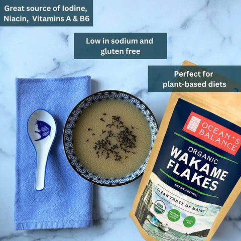 A bowl of soup with Ocean’s Balance Organic Wakame Flakes, a spoon on a blue napkin, and the product pack. Text highlights their plant-based, low-sodium, gluten-free benefits and USDA organic certification.