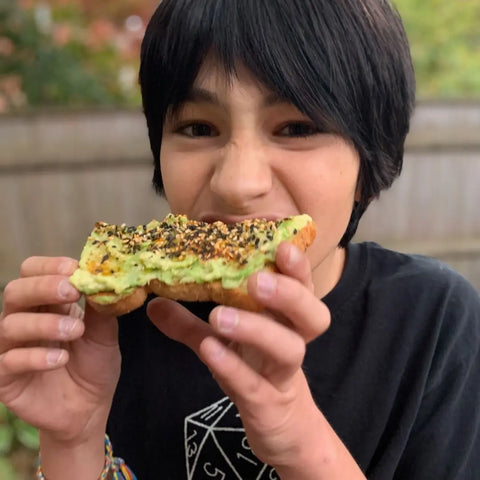 A dark-haired child in a black geometric shirt takes a big bite of avocado toast topped with Oceans Balance Spicy Furikake, outdoors by a blurred wooden fence and greenery.