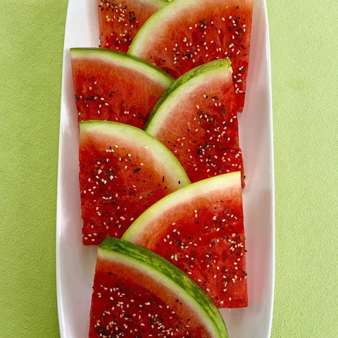 Several triangular watermelon slices with green rind are neatly arranged on a white rectangular plate, sprinkled with Oceans Balance Spicy Furikake made from regeneratively farmed seaweed, on a light green background.