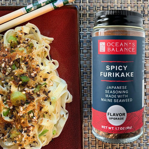 A red plate of seasoned noodles topped with green onions and sesame seeds sits beside a jar of Oceans Balance Spicy Furikake, made with regeneratively farmed Maine seaweed, on a woven placemat with chopsticks.