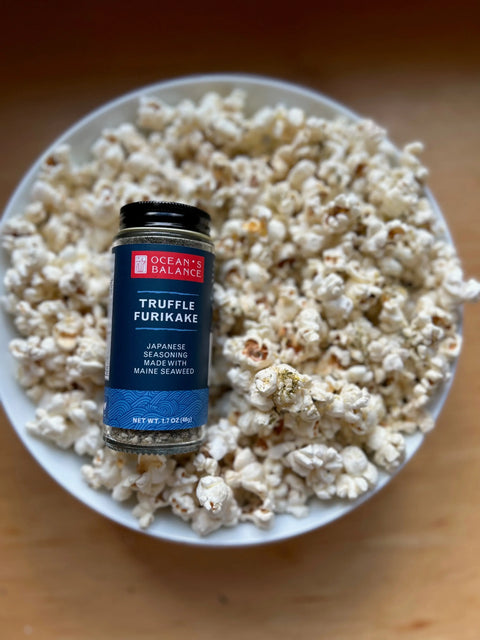 A bottle of Oceans Balance Truffle Furikake, a savory vegan seaweed seasoning, sits atop a bowl of popcorn on a wooden surface—ready to add the perfect touch of Japanese rice seasoning flavor.