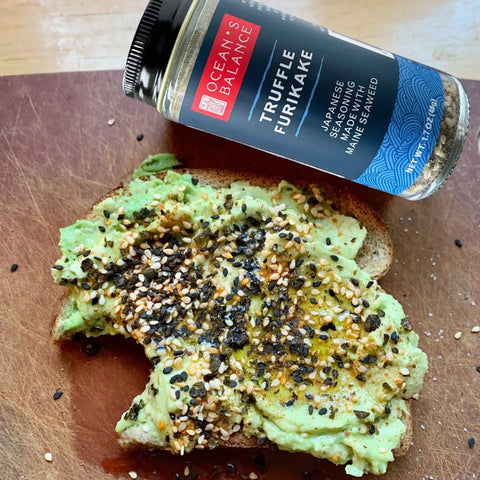 A slice of toast with mashed avocado is sprinkled with Oceans Balance Truffle Furikake, a savory vegan seaweed seasoning, and sits next to its labeled jar on a wooden surface.