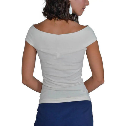 A woman with short, wavy dark hair is seen from the back, wearing the Spiritex | Sustainable Apparel Venus Top in organic cotton with an off-shoulder neckline and a dark blue skirt, one hand on her hip and facing slightly right.