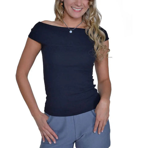 A woman stands smiling against a white background, wearing the Spiritex | Sustainable Apparel Venus Top in black with an off-shoulder neckline and gray shorts, with her hands in her pockets.