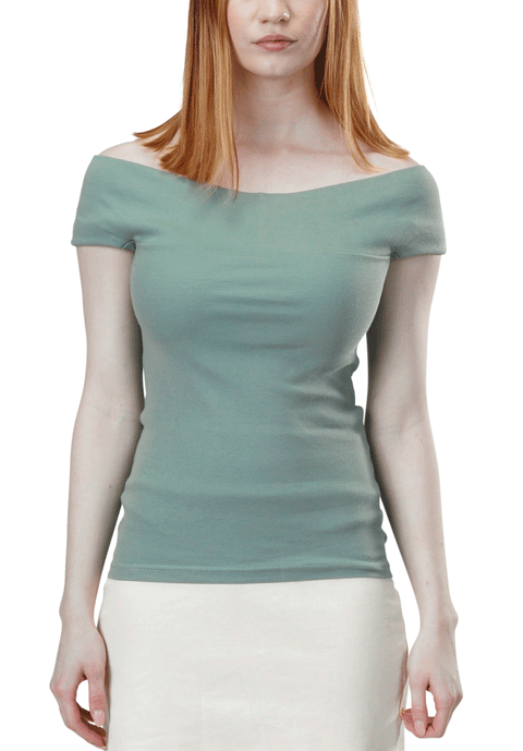 A woman with straight, shoulder-length red hair wears the Spiritex Venus Top—a light green, off-shoulder organic cotton blouse—paired with a white skirt, standing against a white background.