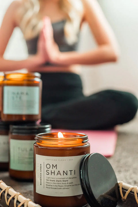 An Om Shanti Amber Soy Candle by Shanti + Tula, USA-made for calm and meditation, glows on a tray as a woman sits cross-legged on a yoga mat, hands in prayer, surrounded by more softly lit candles.