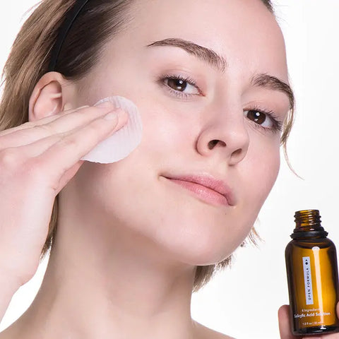 A person with clear skin gently applies Open Formula’s Blemish Control Superset to their cheek using a cotton pad, while holding the brown bottle and appearing calm.