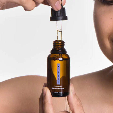 A person dispenses Open Formula’s Blemish Control Superset from an amber bottle with a dropper, highlighting part of their face and shoulder against a plain white background.