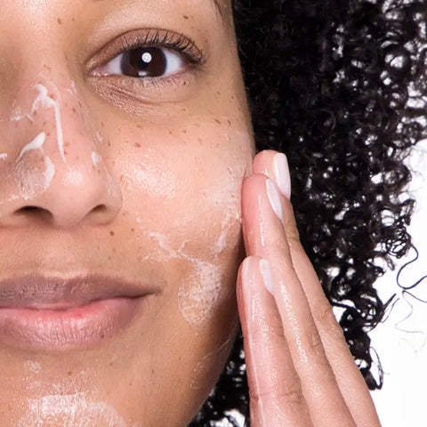 A person with curly hair gently massages Open Formula Gentle Cleanser onto their cheek, creating a foam for deep pore cleansing while maintaining a clear complexion.
