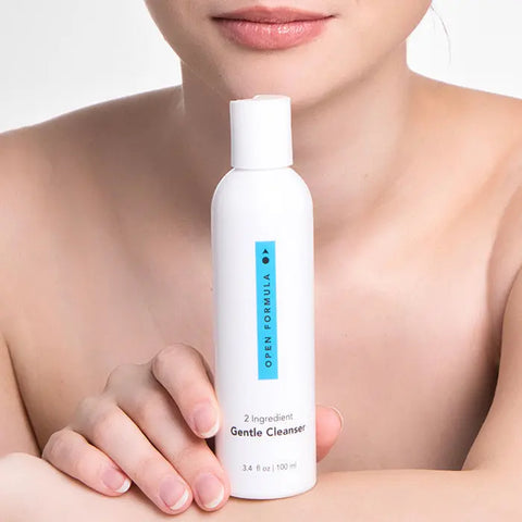 A woman with bare shoulders holds a white Open Formula Gentle Cleanser bottle in front of her chest, showcasing this foaming cleanser for sensitive skin; only the lower half of her face is visible.