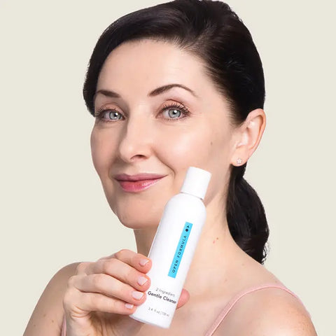 A woman with fair skin and dark hair smiles, holding Open Formulas Gentle Cleanser—a white bottle—close to her face against a plain light background, highlighting its deep pore cleaning benefits.