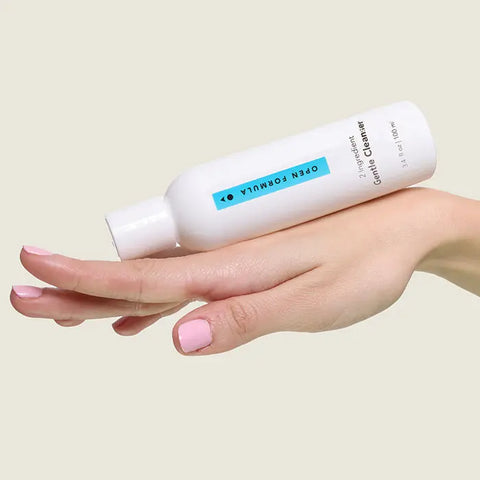 A hand with light pink nail polish holds a white Open Formula Gentle Cleanser bottle—a gentle foaming cleanser for sensitive skin—against a beige background.