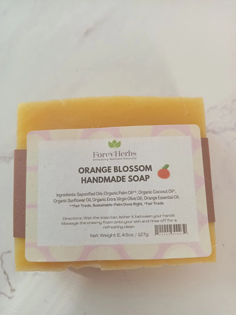 A 4.5oz rectangular bar of Forev'Herbs Orange Blossom Handmade Soap features a white label with an orange icon, the product name, and highlights organic ingredients including orange essential oil.