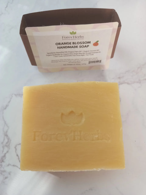 A bar of Forev'Herbs Orange Blossom Handmade Soap sits on marble, featuring its embossed logo. The packaging behind highlights its hydrating lather and natural orange essential oil ingredients.