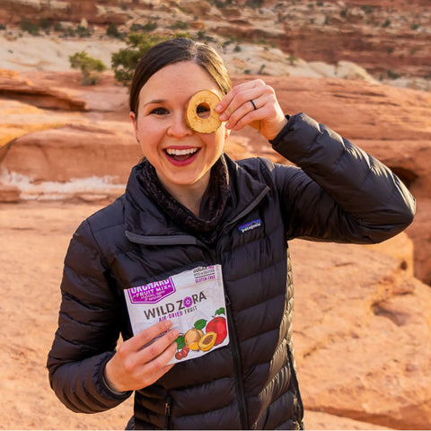 A woman in a black jacket smiles outdoors, holding a dried apple ring from Wild Zoras Fruits - Orchard Air-Dried Mix With Cherries, Apples & Apricots 2-Pack over one eye and the pack in her other hand, with stunning red rocks behind her.