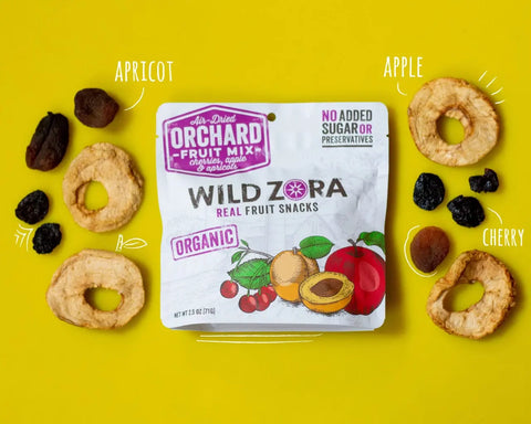 A Wild Zora Fruits - Orchard Air-Dried Mix With Cherries, Apples & Apricots 2-Pack on a yellow background, surrounded by organic apples, apricots, and cherries with handwritten labels for each dried fruit.