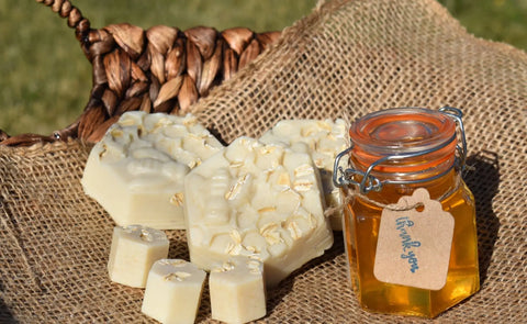 A jar of honey with a thank you tag sits on burlap next to Bathsheba Body Works Body Butter Scrub - Country Butter, handmade soaps topped with oats, and an exfoliating scrub, with a woven basket and grass in the background.