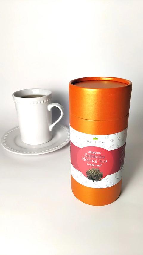 A white teacup and saucer sit beside an orange cylindrical container labeled “Forev'Herbs Organic Bulukutu Herbal Tea,” showcasing this aromatic herbal tea blend, inspired by traditional African teas, on a pristine white surface.